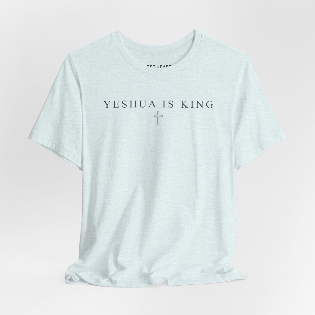 Pale heather ice blue t-shirt with small YESHUA IS KING text above a thin outlined cross, shown front on a light gray background