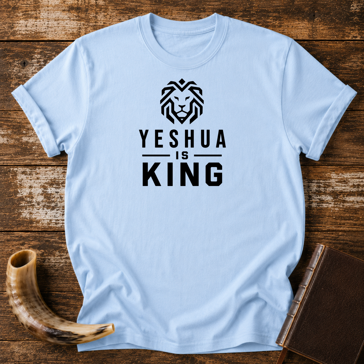 Light blue t-shirt with a geometric lion head above YESHUA IS KING lettering, laid flat with a shofar and a leather book