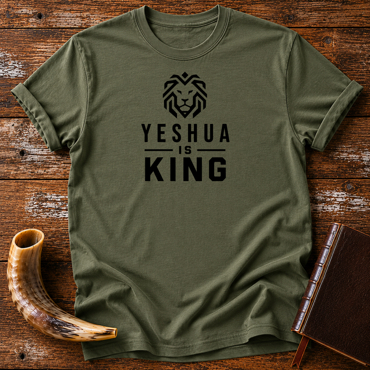 Military green t-shirt with a geometric lion head above YESHUA IS KING lettering, laid flat with a shofar and a leather book