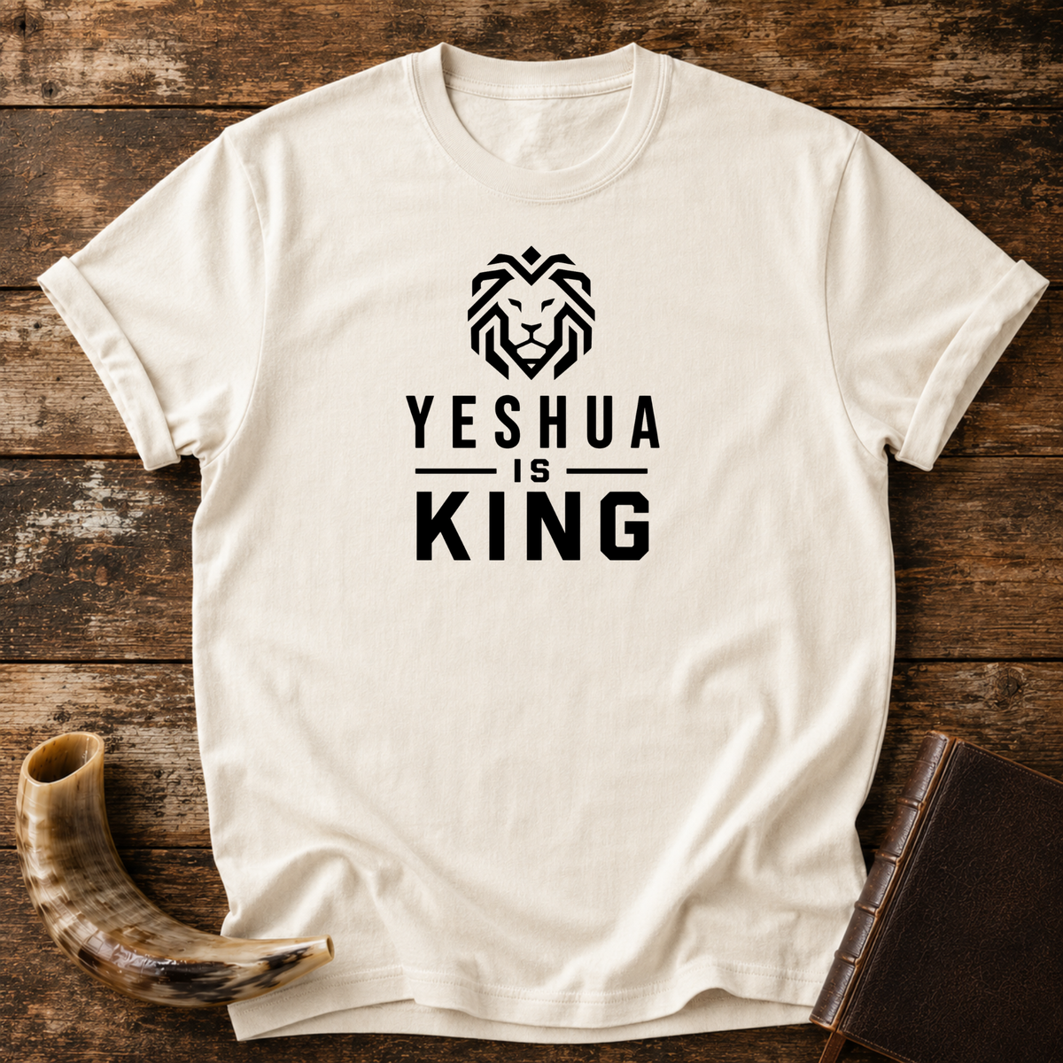 Natural cream t-shirt with a geometric lion head above YESHUA IS KING lettering, laid flat with a shofar and a leather book