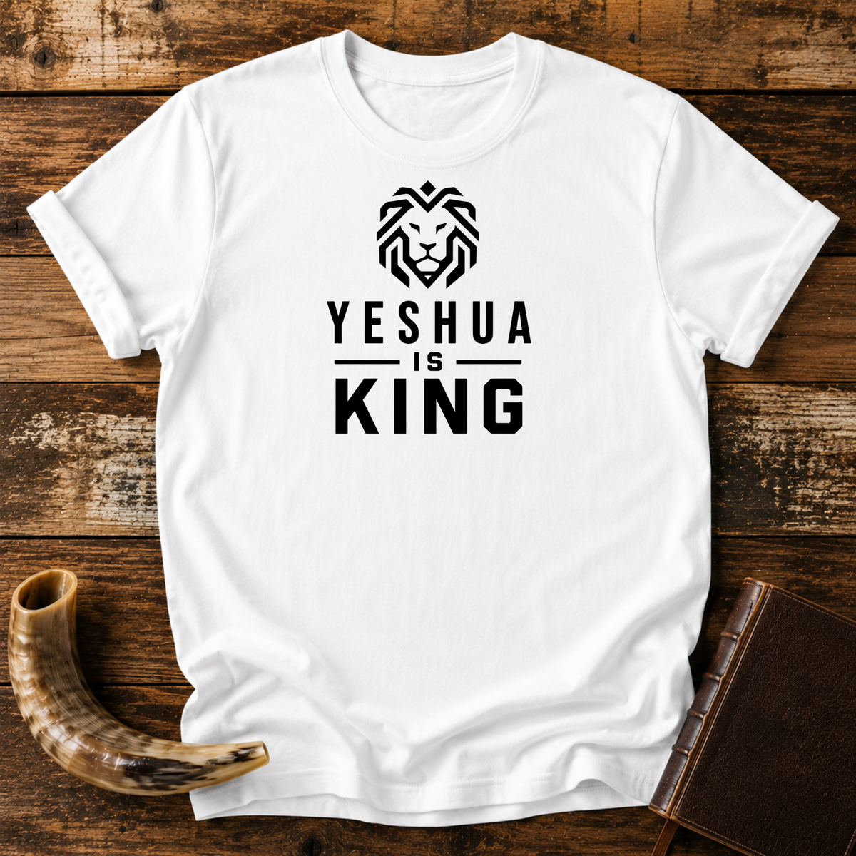 White t-shirt with a geometric lion head above YESHUA IS KING lettering, laid flat with a shofar and a leather book