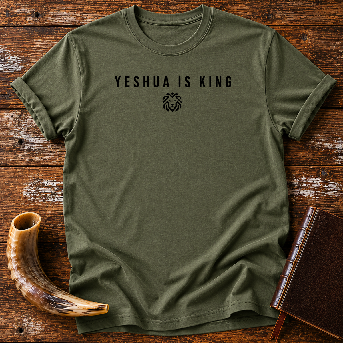 Military green t-shirt with small YESHUA IS KING text and a tiny lion emblem across the chest, laid flat with a shofar and a leather book