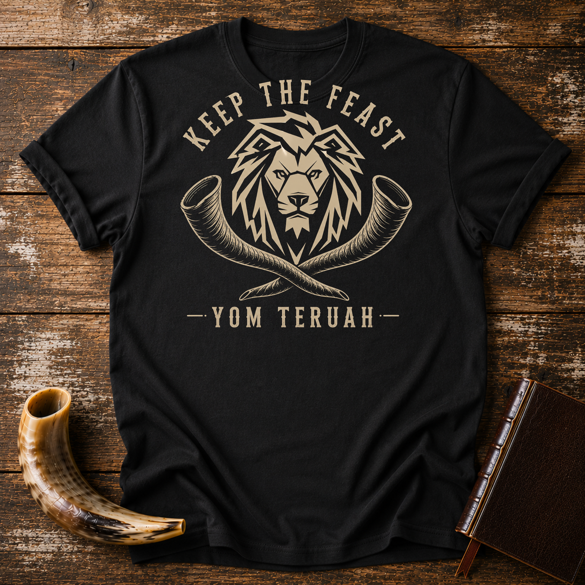 Black t-shirt with a tan lion head over crossed shofars, KEEP THE FEAST YOM TERUAH, laid flat with a shofar and a leather book