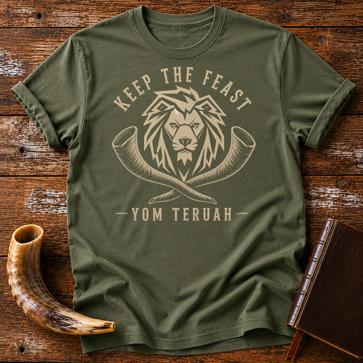 Military green t-shirt with a tan lion head over crossed shofars, KEEP THE FEAST YOM TERUAH, laid flat with a shofar and a leather book