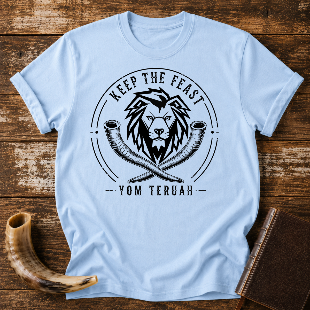 Light blue t-shirt with a black lion head over crossed shofars, KEEP THE FEAST YOM TERUAH, laid flat with a shofar and a leather book