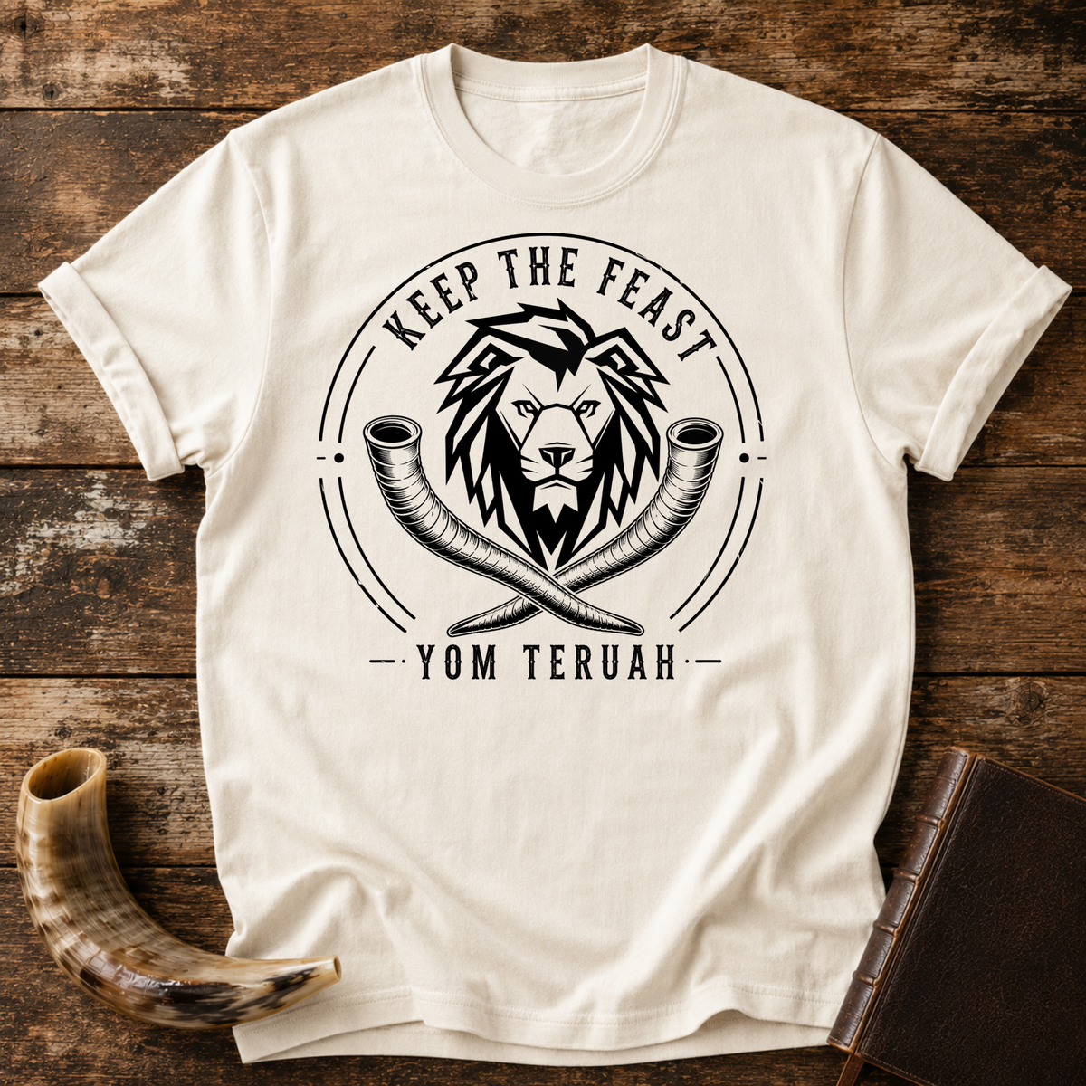 Natural cream t-shirt with a black lion head over crossed shofars, KEEP THE FEAST YOM TERUAH, laid flat with a shofar and a leather book